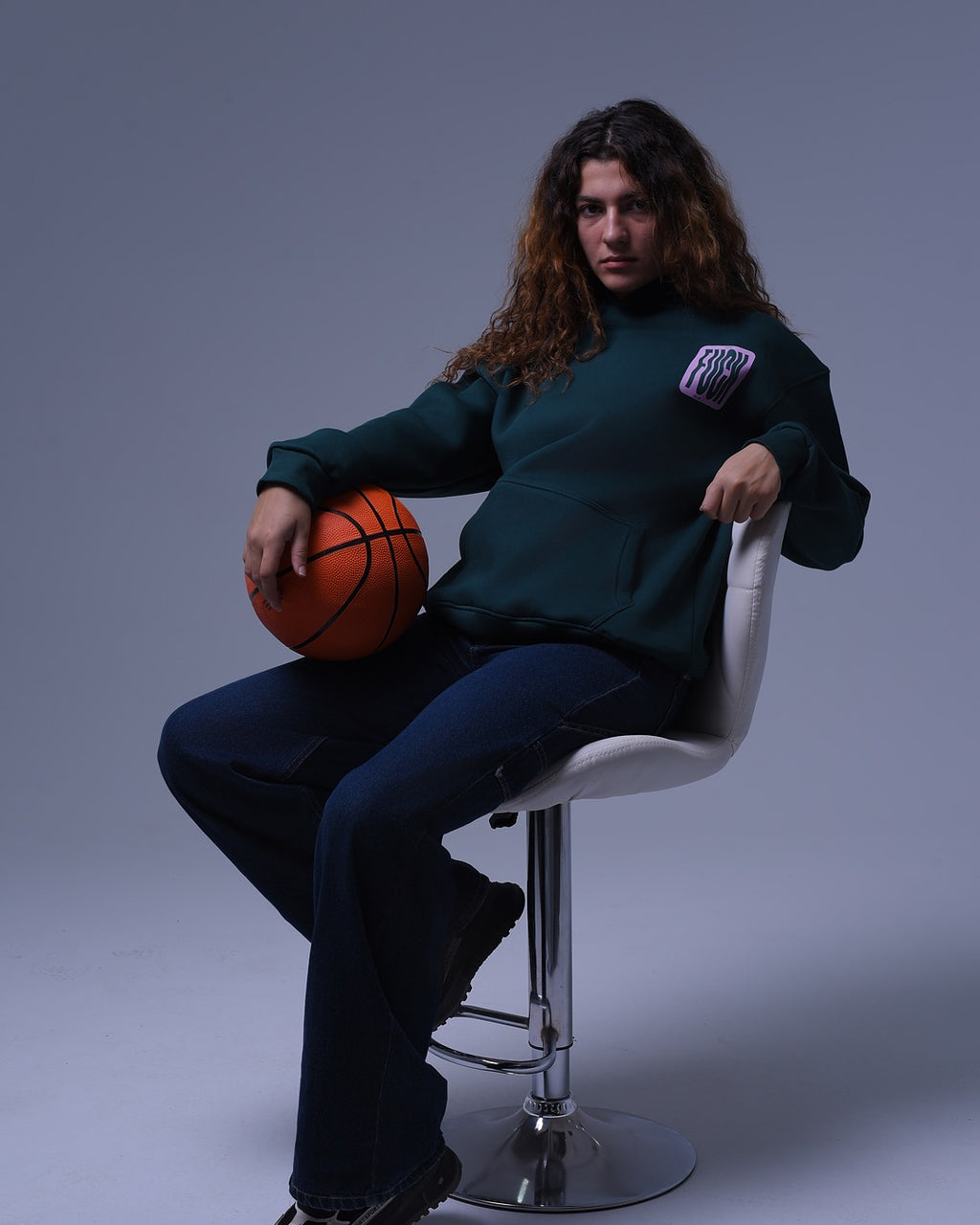 NotYourSize2025 oversized hoodie for unisex in dark green worn by model holding a basketball seated on a white stool