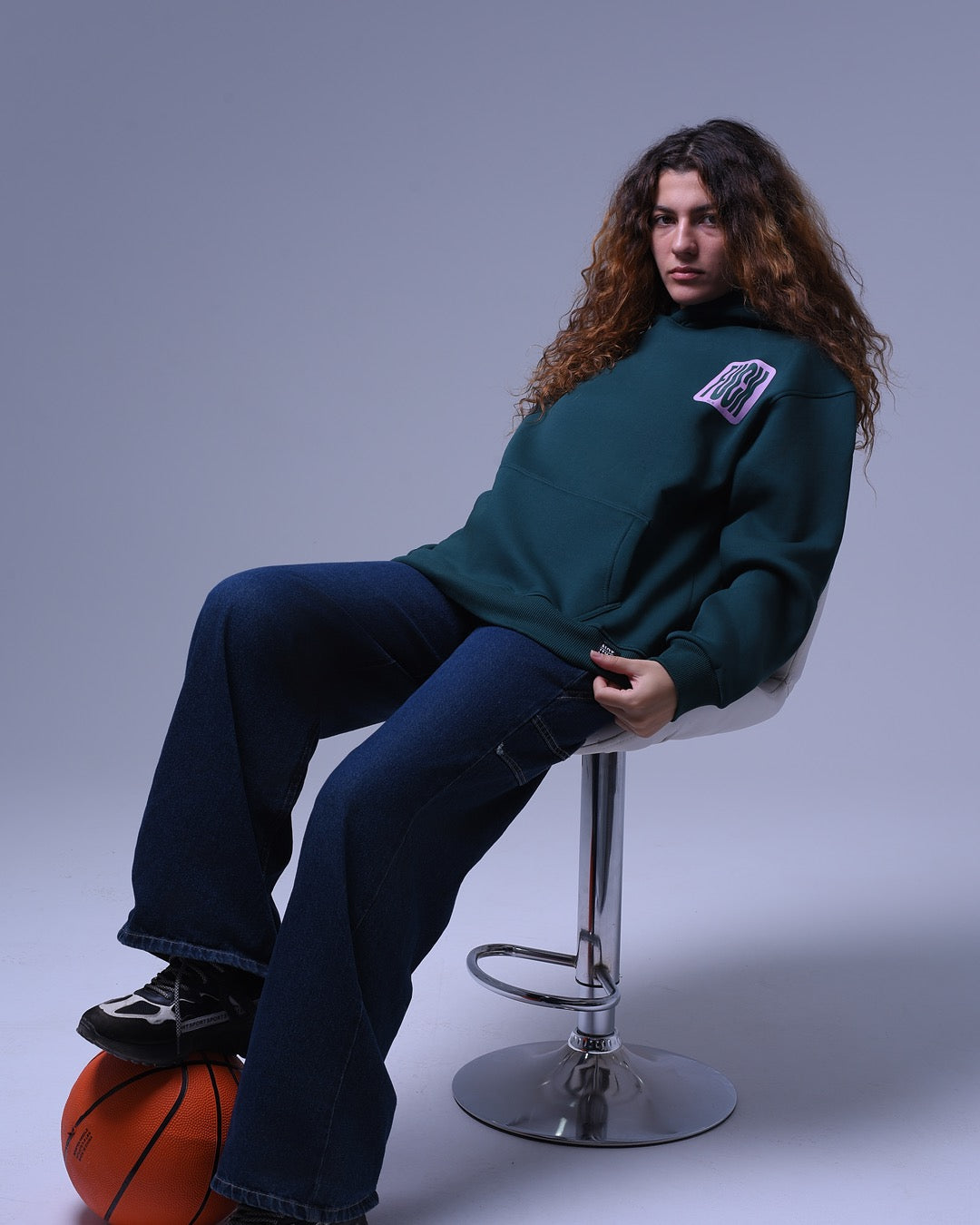 Person wearing oversized hoodie for unisex in dark green sitting on a stool with basketball