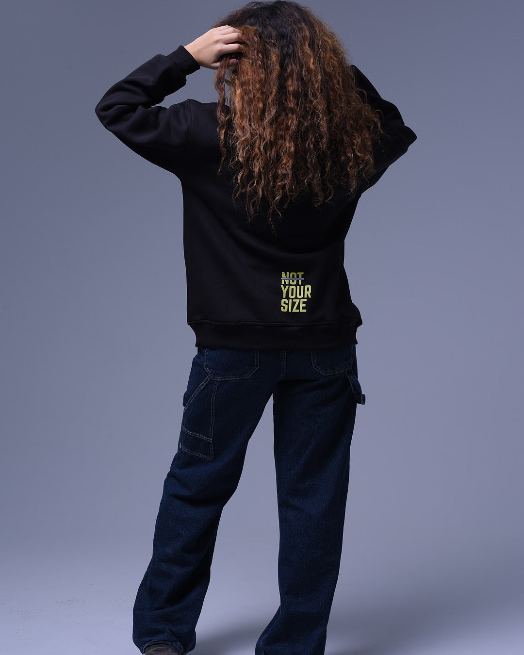 Back view of person wearing black oversized crewneck sweatshirt with NotYourSize2025 logo on lower back